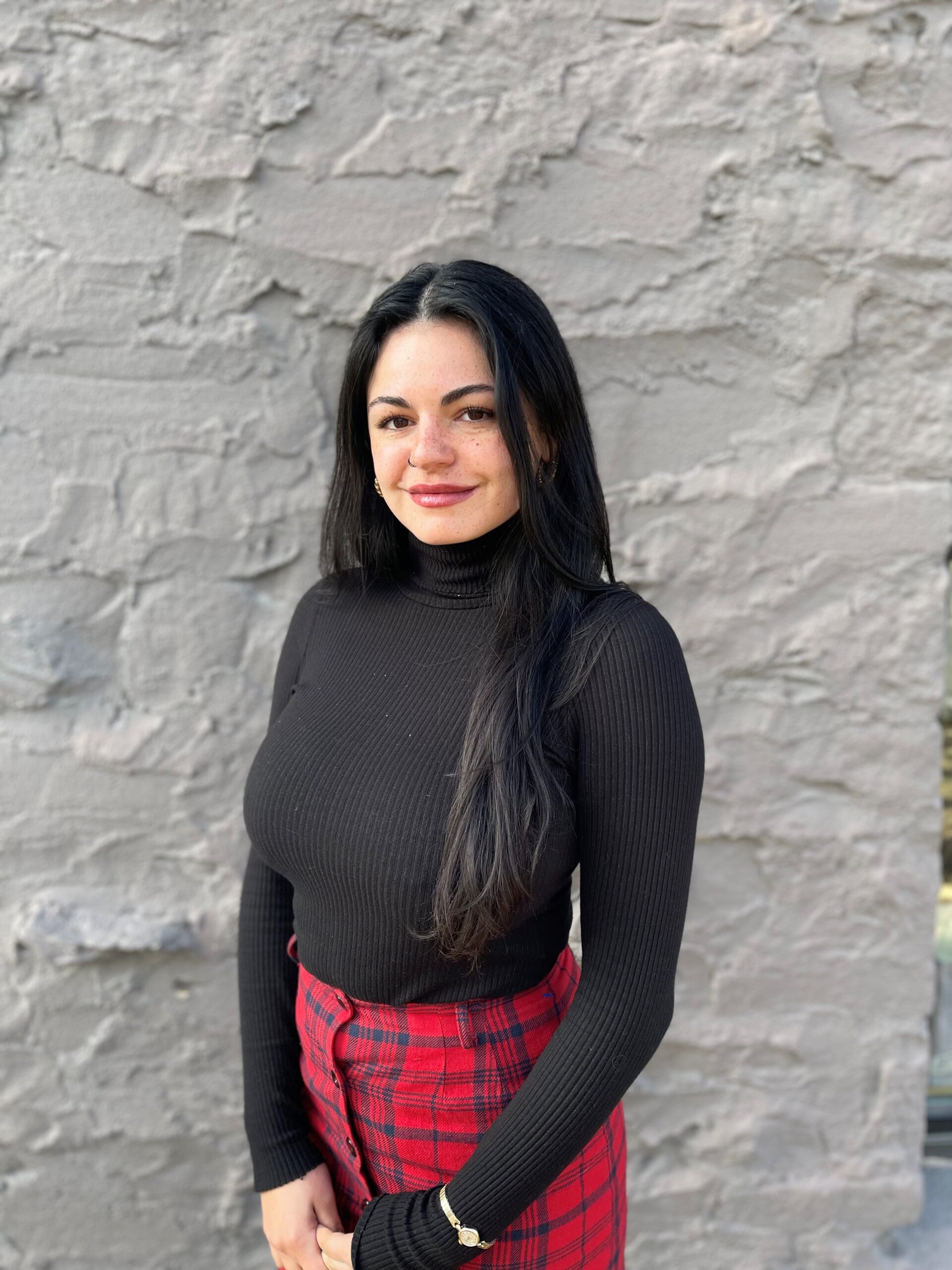 A women in black turtle neck and red plaid pants with long black hair poses against a grey background.