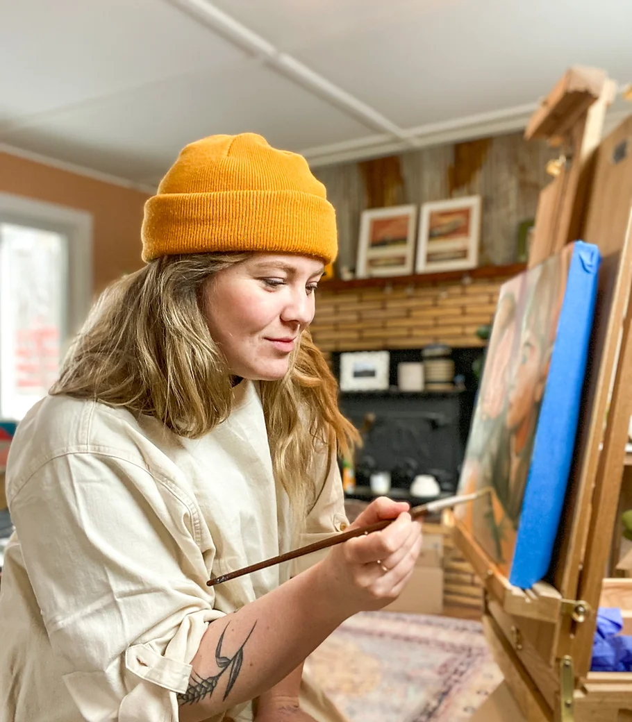 An artist poses with paintbrush in hand. She is in her studio space. The artist is named Elise Perpignano.