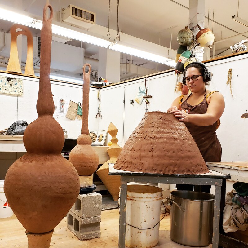 Artist Danielle O'Malley (she/they) works on a large scale clay piece. Next to her are two finished pieces.